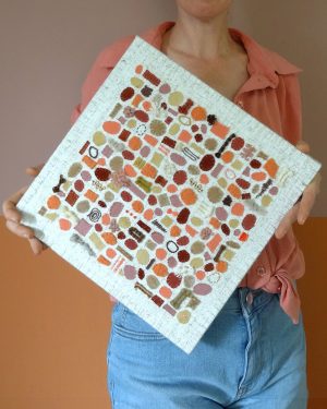 Dit werk heet Avondzon en zachte perzik en is 30x30 cm. Avondzon en zachte perzik brengt stoffen met een persoonlijk verhaal samen in een tactiel kunstwerk vol warme kleuren en verschillende texturen Annemieke heeft het werk schuin in haar handen. Het matcht goed met haar roze bloes.