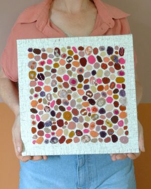 Dit werk heet Pluktuin in bloei en is 30x30 cm. Pluktuin in bloei is een speels en kleurrijk werk vol persoonlijke stoffen en herinneringen. Annemieke houdt het werk in haar handen.