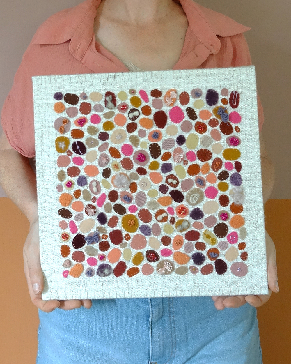 Dit werk heet Pluktuin in bloei en is 30x30 cm. Pluktuin in bloei is een speels en kleurrijk werk vol persoonlijke stoffen en herinneringen. Annemieke houdt het werk in haar handen.