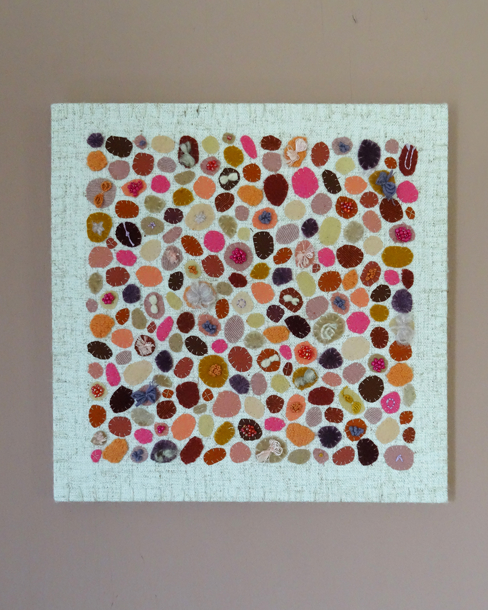 Dit werk heet Pluktuin in bloei en is 30x30 cm. Pluktuin in bloei is een speels en kleurrijk werk vol persoonlijke stoffen en herinneringen. Dit is de voorkant van het werk. Het zijn allemaal ronde vormen van verschillende soorten stof afgewerkt met kralen.