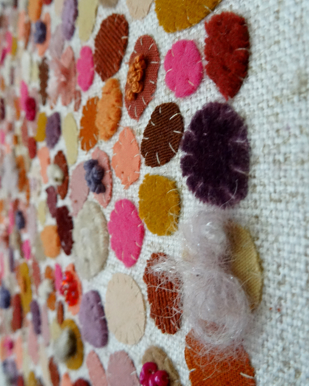 Dit werk heet Pluktuin in bloei en is 30x30 cm. Pluktuin in bloei is een speels en kleurrijk werk vol persoonlijke stoffen en herinneringen. Hier zie je een close-up van het werk. Het bestaat uit allemaal ronde vormen waarbij een paars fluffie wol opvalt.