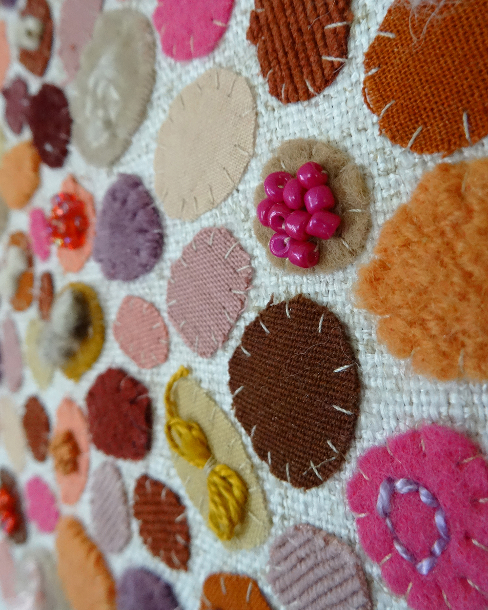 Dit werk heet Pluktuin in bloei en is 30x30 cm. Pluktuin in bloei is een speels en kleurrijk werk vol persoonlijke stoffen en herinneringen. Hier zie je een close-up van het werk. Het bestaat uit allemaal ronde vormen waarbij een cluster van roze kraaltjes opvalt.