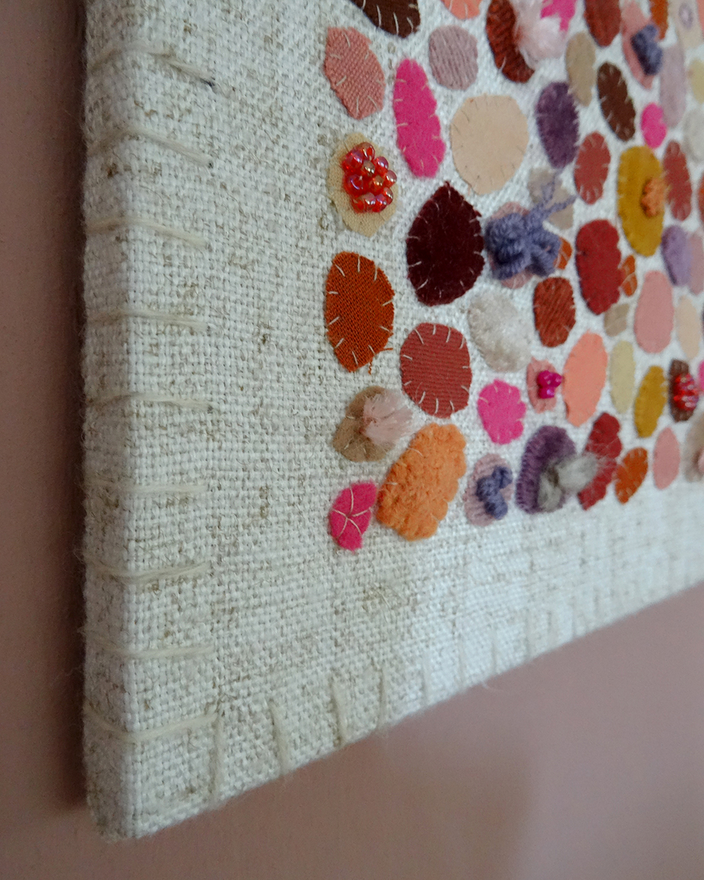 Dit werk heet Pluktuin in bloei en is 30x30 cm. Pluktuin in bloei is een speels en kleurrijk werk vol persoonlijke stoffen en herinneringen. Hier zie je de linker onderhoek van het werk. Het bestaat uit allemaal ronde vormen gemaakt van verschillende soorten stoffen en kralen.