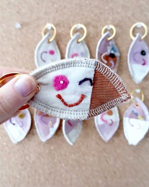 Voorkant van een handgemaakte sleutelhanger van hergebruikte stoffen. De sleutelhanger heeft een zachte textiele afwerking en maakt deel uit van de serie Knipoogjes. De voorkant is roze katoen met oranje ribstof. Hierop is een knipoog gezicht geborduurd met diverse kleurtjes. Dit is knipoog nummer 1.