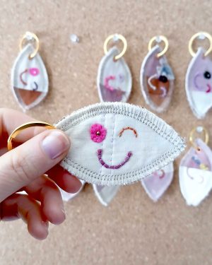 Voorkant van een handgemaakte sleutelhanger van hergebruikte stoffen. De sleutelhanger heeft een zachte textiele afwerking en maakt deel uit van de serie Knipoogjes. De voorkant is gemaakt van egaal roze katoen. Hierop is een knipoog gezicht geborduurd met diverse kleurtjes. Dit is knipoog nummer 2.