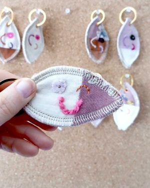 Voorkant van een handgemaakte sleutelhanger van hergebruikte stoffen. De sleutelhanger heeft een zachte textiele afwerking en maakt deel uit van de serie Knipoogjes. De voorkant is roze katoen en een klein stukje roze met bloemetjes. Hierop is een knipoog gezicht geborduurd met diverse kleurtjes. Dit is knipoog nummer 3.