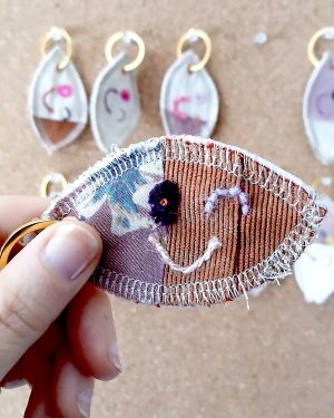 Voorkant van een handgemaakte sleutelhanger van hergebruikte stoffen. De sleutelhanger heeft een zachte textiele afwerking en maakt deel uit van de serie Knipoogjes. De voorkant is gemaakt van oranje ribstof en een deel roze bloemetjesstof. Hierop is een knipoog gezicht geborduurd met diverse kleurtjes. Dit is knipoog nummer 4.