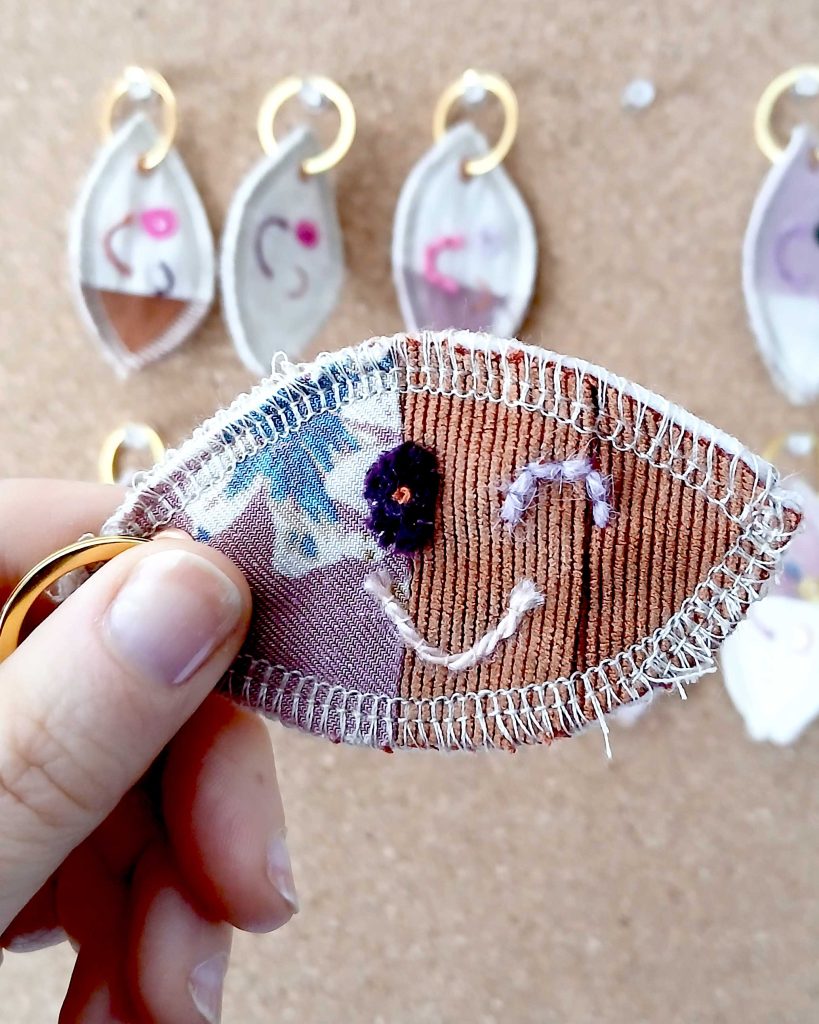 Voorkant van een handgemaakte sleutelhanger van hergebruikte stoffen. De sleutelhanger heeft een zachte textiele afwerking en maakt deel uit van de serie Knipoogjes. De voorkant is gemaakt van oranje ribstof en een deel roze bloemetjesstof. Hierop is een knipoog gezicht geborduurd met diverse kleurtjes. Dit is knipoog nummer 4.