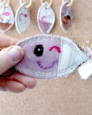 Voorkant van een handgemaakte sleutelhanger van hergebruikte stoffen. De sleutelhanger heeft een zachte textiele afwerking en maakt deel uit van de serie Knipoogjes. De voorkant is gemaakt van roze katoen met roze ribstof. Hierop is een knipoog gezicht geborduurd met diverse kleurtjes. Dit is knipoog nummer 5.