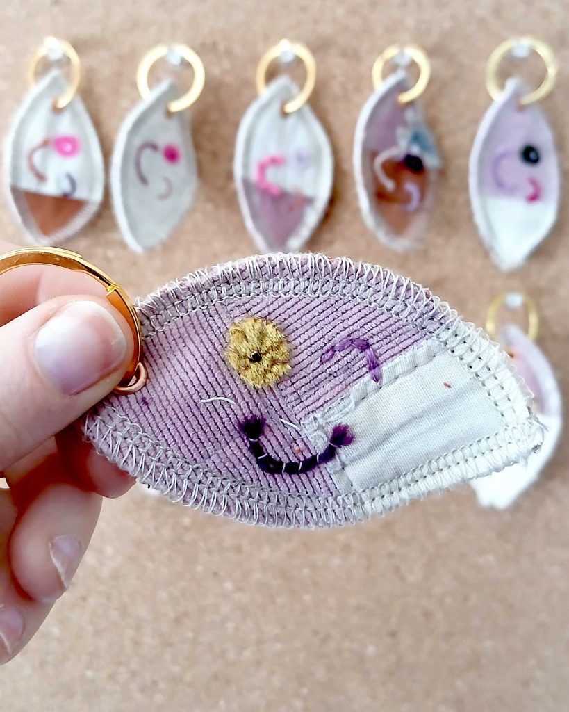 Voorkant van een handgemaakte sleutelhanger van hergebruikte stoffen. De sleutelhanger heeft een zachte textiele afwerking en maakt deel uit van de serie Knipoogjes. De voorkant is gemaakt van roze ribstof met een klein puntje egale roze katoen. Hierop is een knipoog gezicht geborduurd met diverse kleurtjes. Dit is knipoog nummer 6.