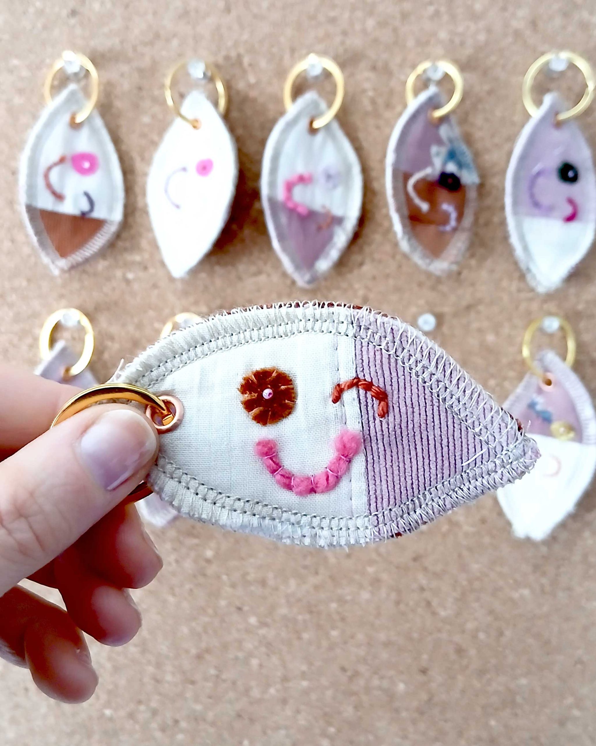 Voorkant van een handgemaakte sleutelhanger van hergebruikte stoffen. De sleutelhanger heeft een zachte textiele afwerking en maakt deel uit van de serie Knipoogjes. De voorkant is gemaakt van roze katoen met roze ribstof. Hierop is een knipoog gezicht geborduurd met diverse kleurtjes. Dit is knipoog nummer 9.